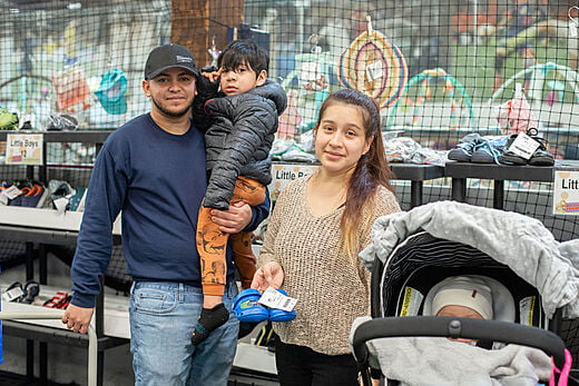 Father holding his son standing next to the mother with her hand on the stroller. There is a baby in the stroller. They are all in front of a table with baby items for sale.