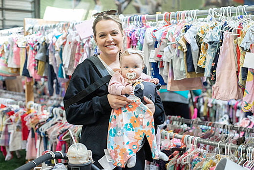 A woman carries a baby in a front carrier. The woman is standing in front of a rack of childrens clothing.